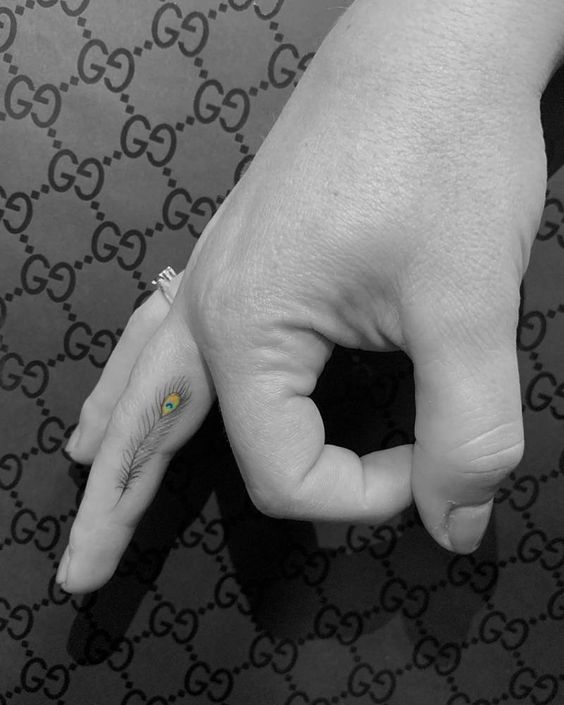 Small and delicate peacock feather tattoo on the side of the finger, featuring fine black linework with subtle green and blue detailing.