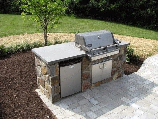 Built-in grill with fridge and cabinets on a paved patio, surrounded by lush grass and vibrant plants