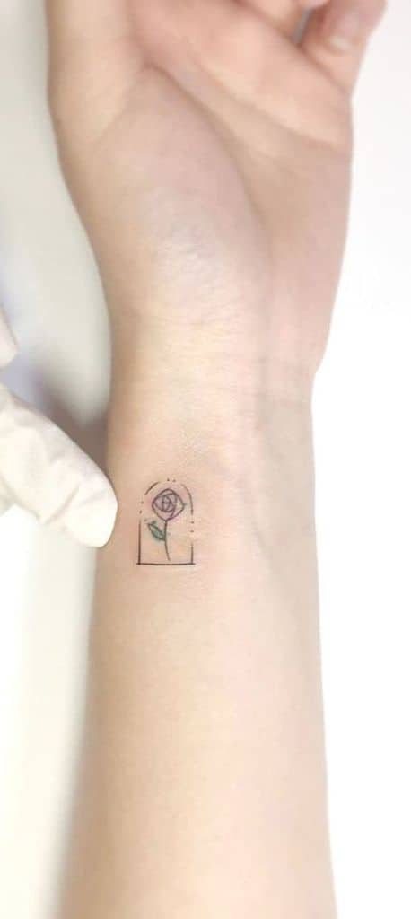 Tiny minimalist tattoo of the enchanted rose inside a glass dome, placed delicately on the wrist with fine-line detailing.