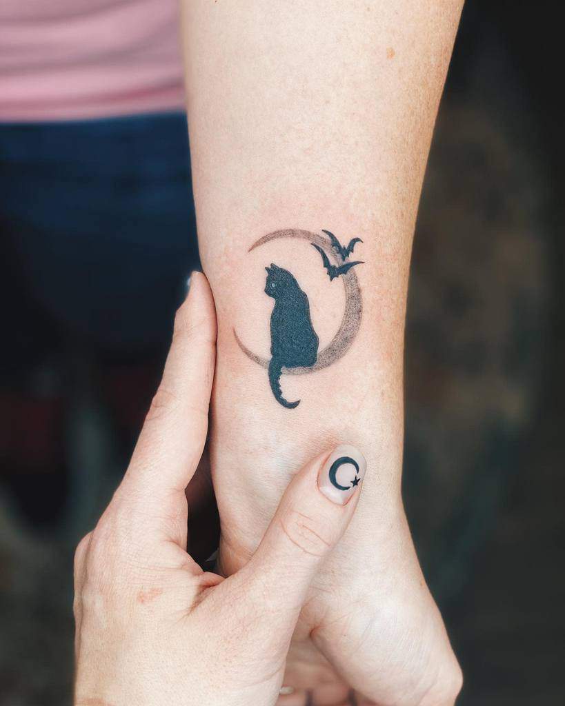 Tattoo of a black cat and bat inside a crescent moon on an inner forearm