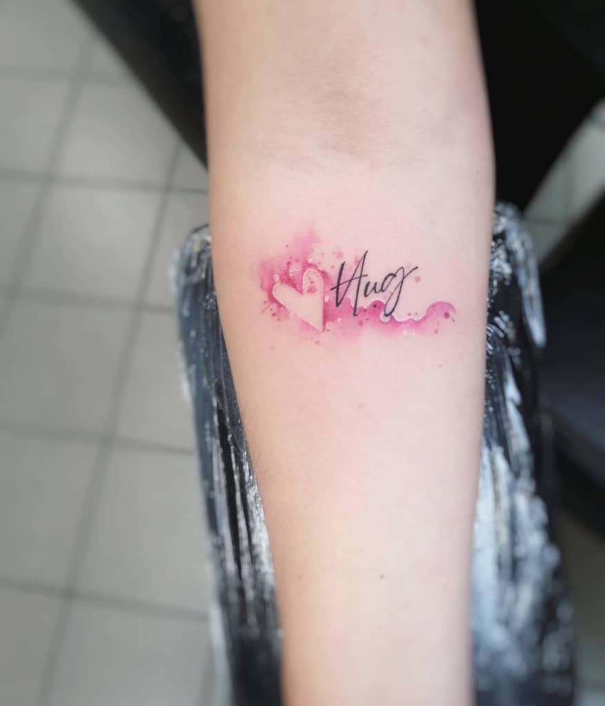 Forearm tattoo with a pink heart and "Hug" written in cursive, surrounded by watercolor splashes