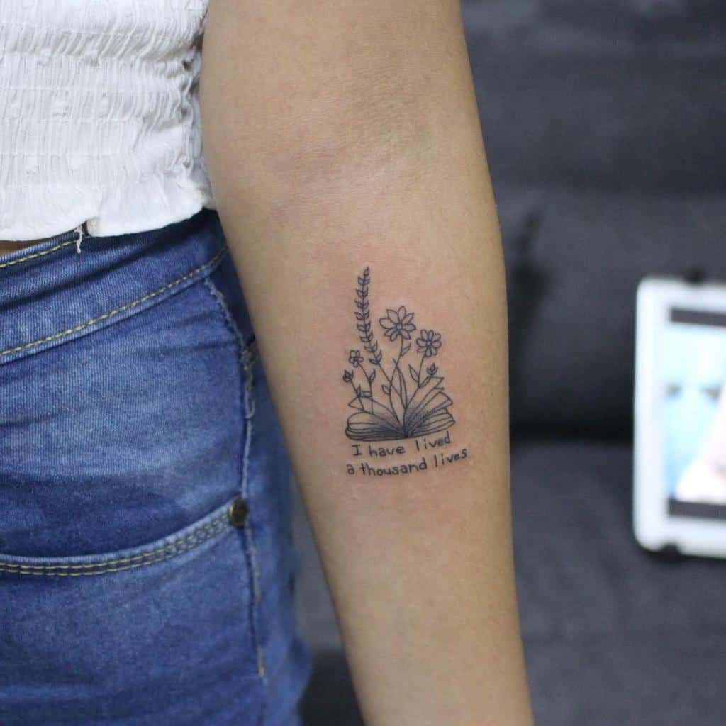 An arm tattoo featuring an open book with delicate flowers intertwined around it and the text 'I have lived a thousand lives' gracefully inscribed below