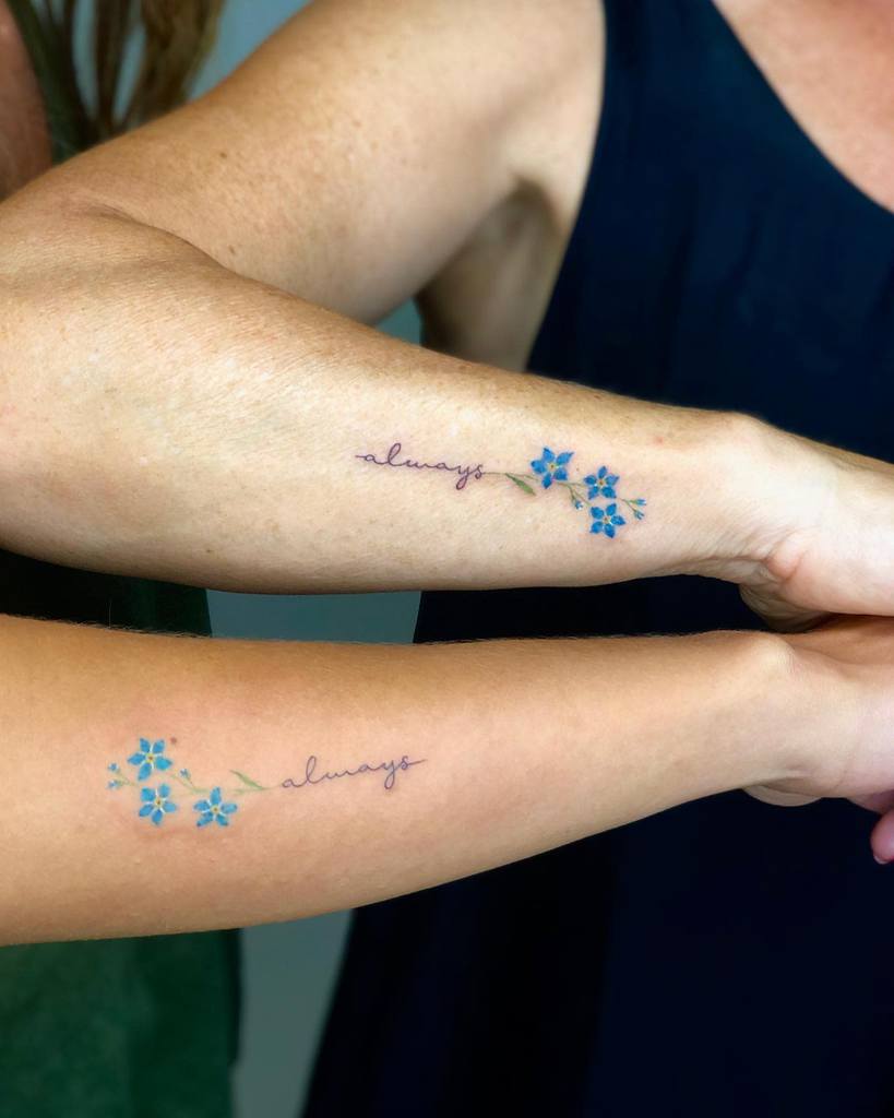 A pair of delicate "always" tattoos, each accompanied by a small bouquet of blue forget-me-nots, inked on the forearms of two people