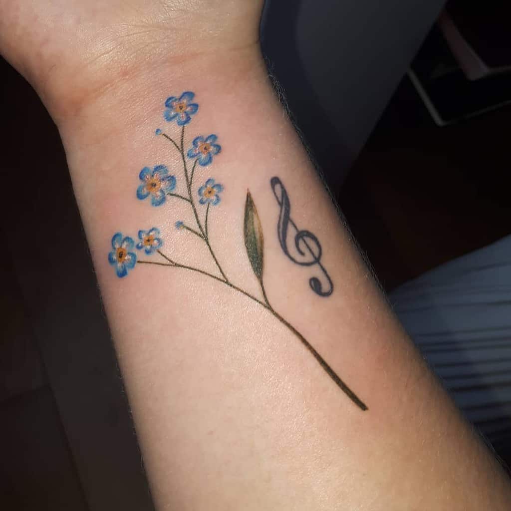 Tattoo of delicate blue forget-me-not flowers intertwined with a musical treble clef on a forearm