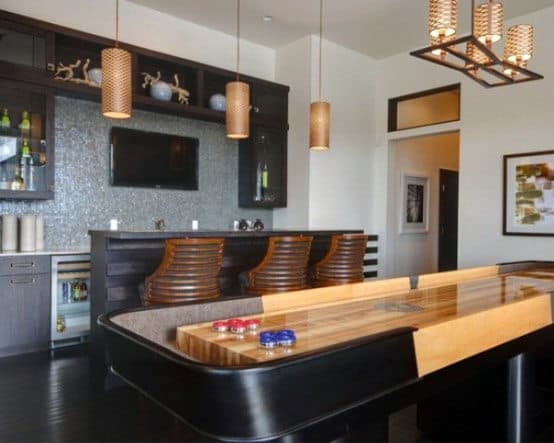 Stylish home bar with a shuffleboard table, wooden bar stools, and hanging pendant lights