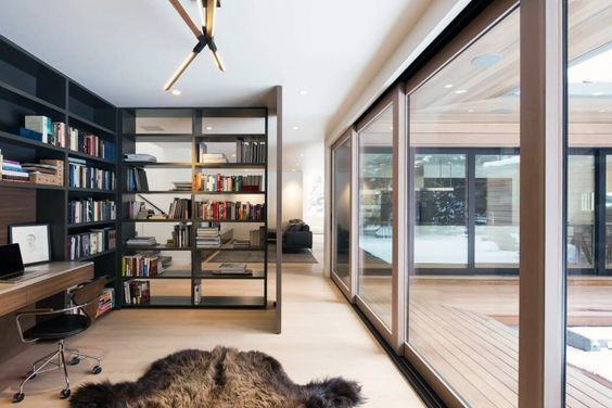 Modern home office with bookshelves, a desk, chair, and large windows overlooking a wooden deck and snowy exterior