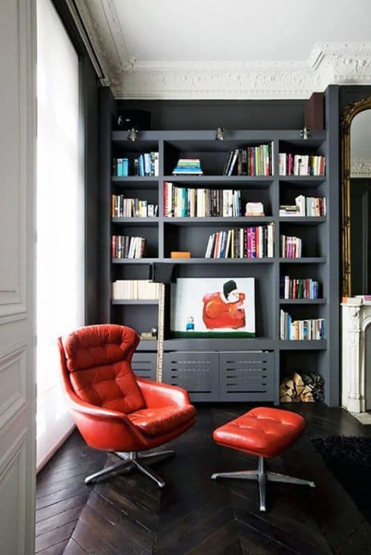 A cozy room with a red armchair and ottoman, bookshelves, a large window, and a painting on display