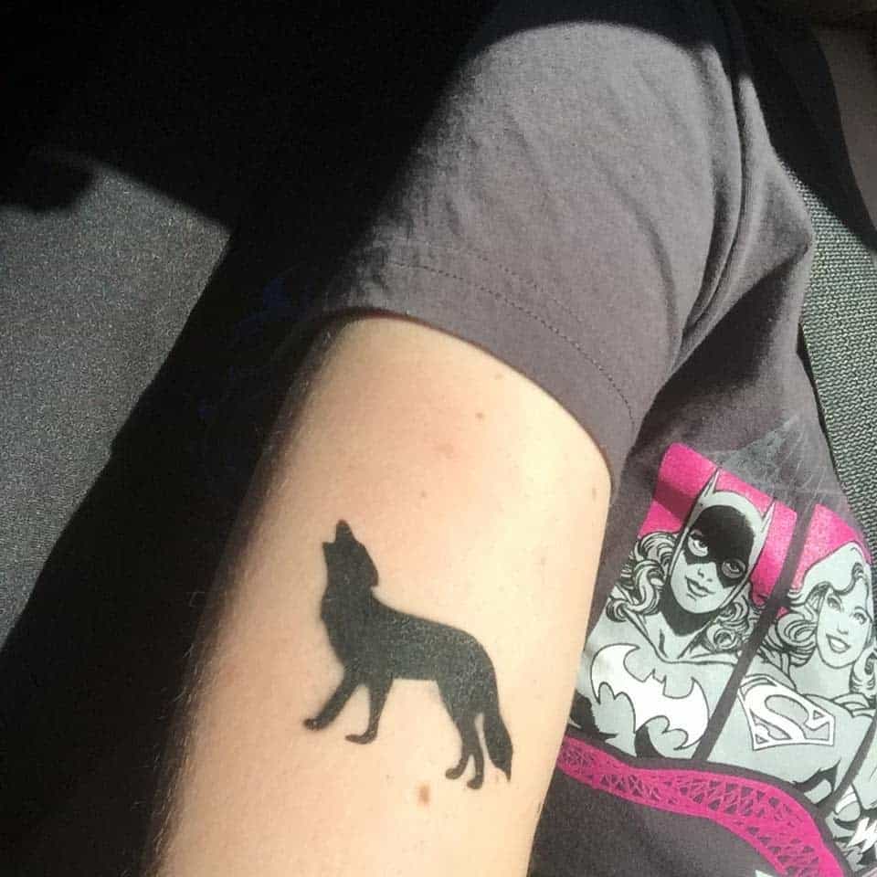 Minimalist black silhouette tattoo of a howling wolf on the upper arm