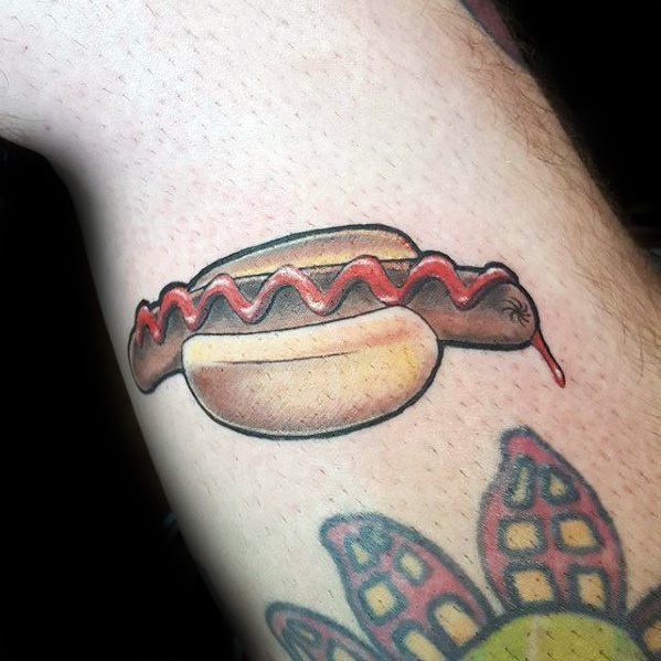 Tattoo of a hot dog with ketchup dripping from the sausage, placed in a bun, on the forearm.
