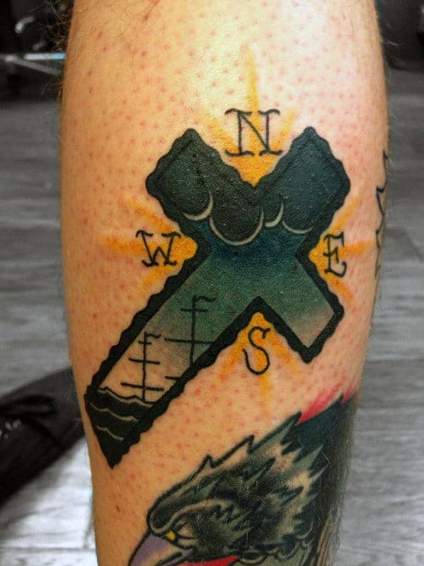 Tattoo of a dark green cross with a wave and staircase design, surrounded by a compass rose showing N, S, E, W, with a yellow glow in the background.
