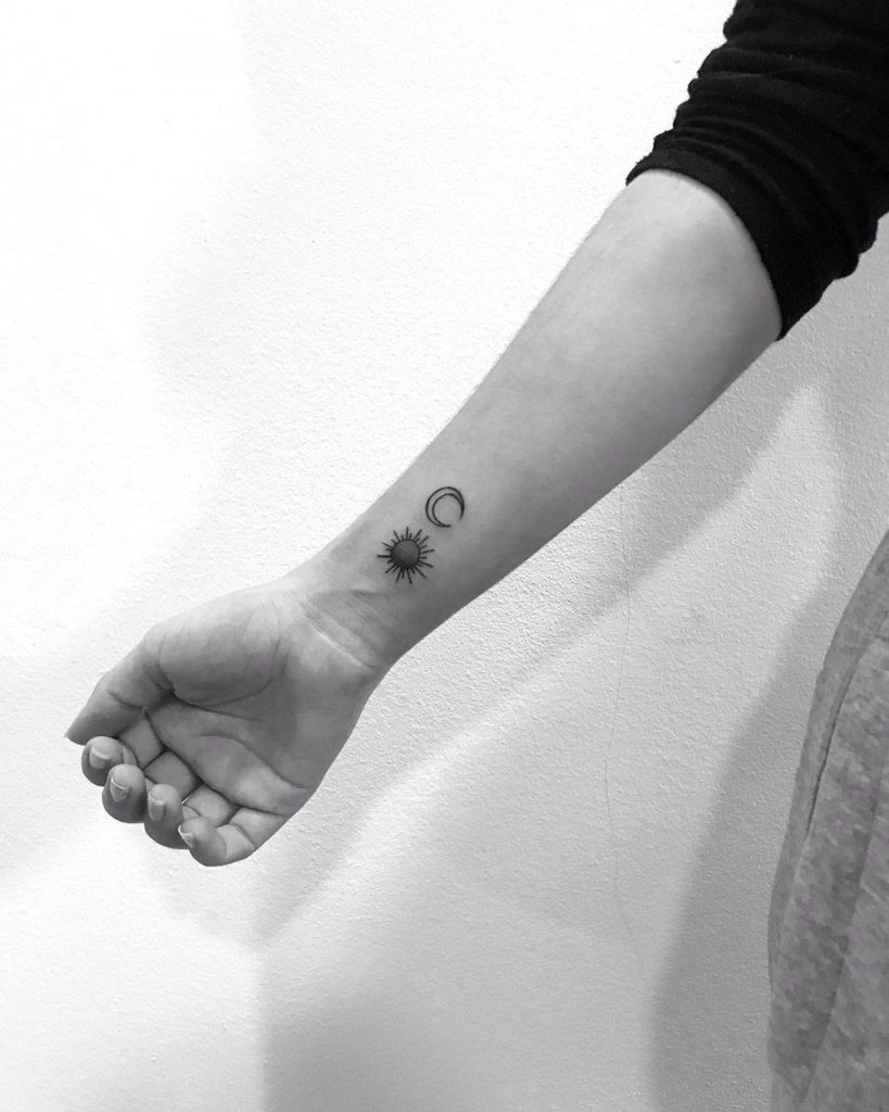 Minimalist black ink tattoo of a small sun and crescent moon on the wrist, featuring fine linework and simple celestial symbols.