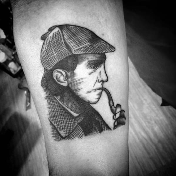 Profile tattoo of Sherlock Holmes in a hat, smoking a pipe, depicted in black and gray tones on an arm