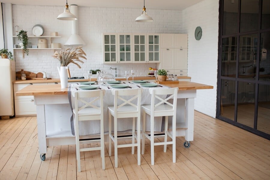 A cozy kitchen featuring a wooden island, three white chairs, pendant lights, and white cabinetry