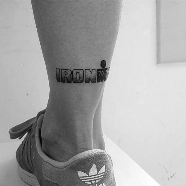 Black ankle tattoo reading IRONMAN 70.3 with the “M-dot” logo replacing the last “A” and a bold, minimalist design style.