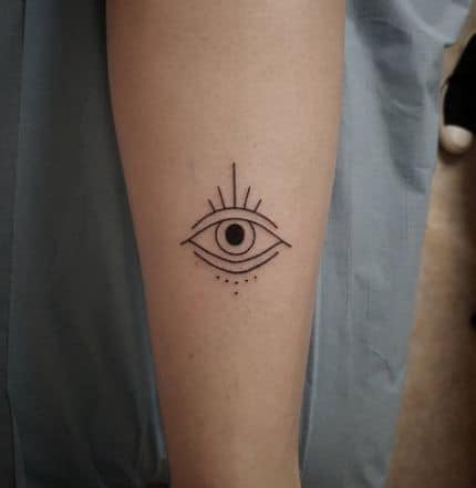 Minimalist tattoo of an all-seeing eye with radiating lines and small dots, inked in fine black lines on the forearm.