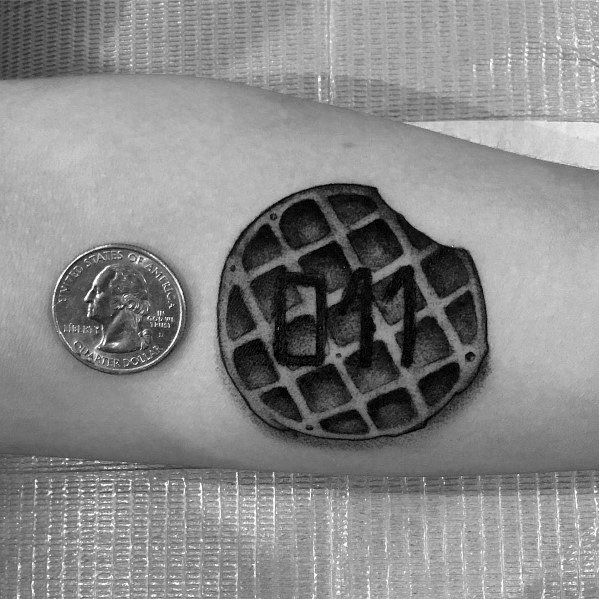 Small black and gray waffle tattoo with a bitten edge and "011" in the center, inspired by pop culture, placed beside a quarter for scale.