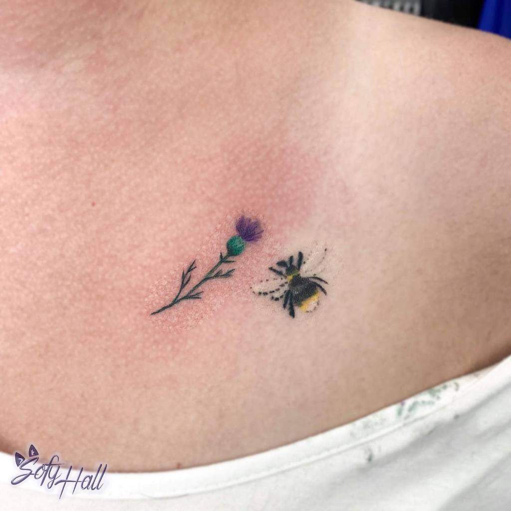 Small and colorful tattoo of a purple thistle and a buzzing bee on the upper chest, showcasing nature's harmony