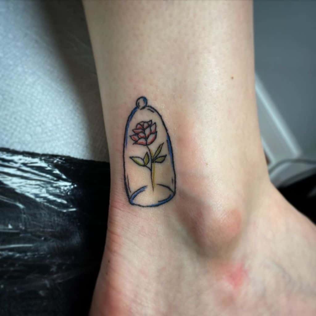 A small, minimalist tattoo of the Beauty and the Beast rose inside a glass dome, with delicate linework and soft red accents on the rose