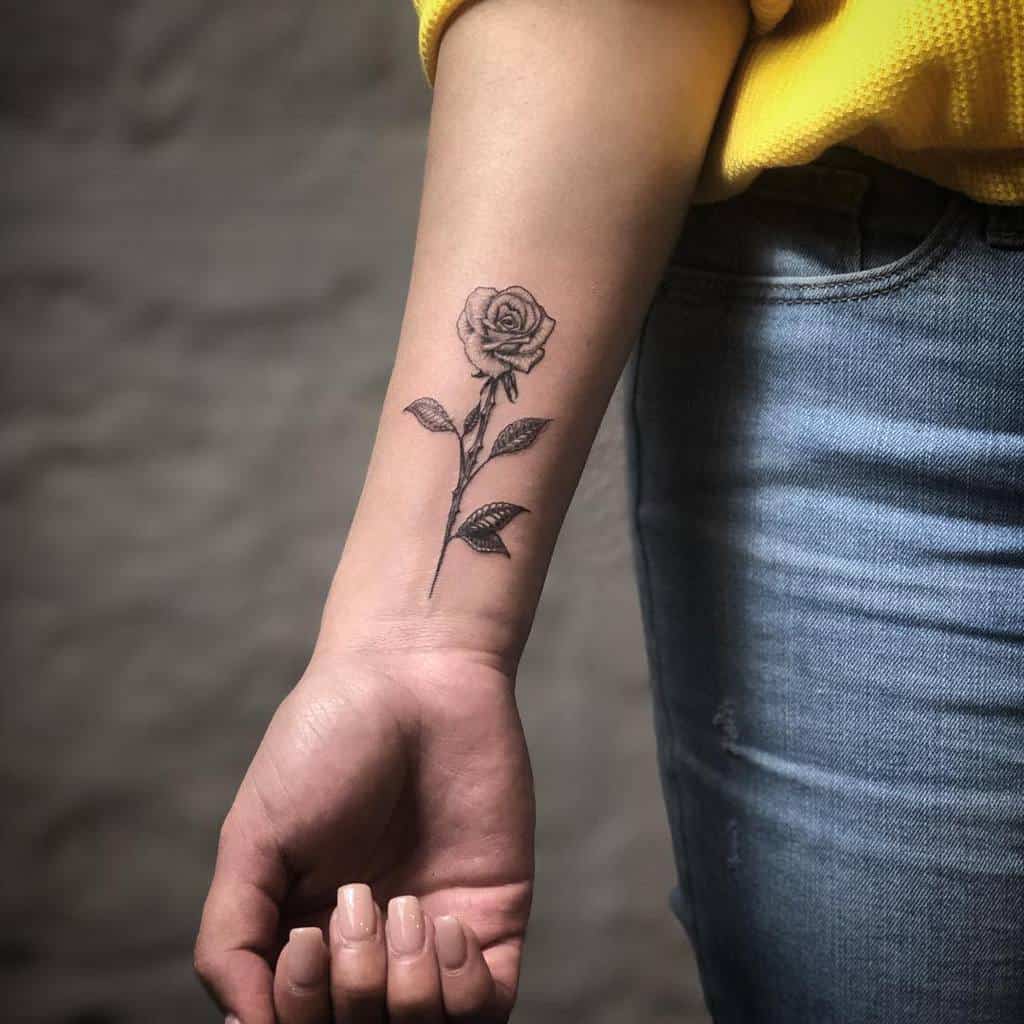 Minimalist black and grey rose tattoo on the forearm, featuring fine linework and a delicate stem with leaves