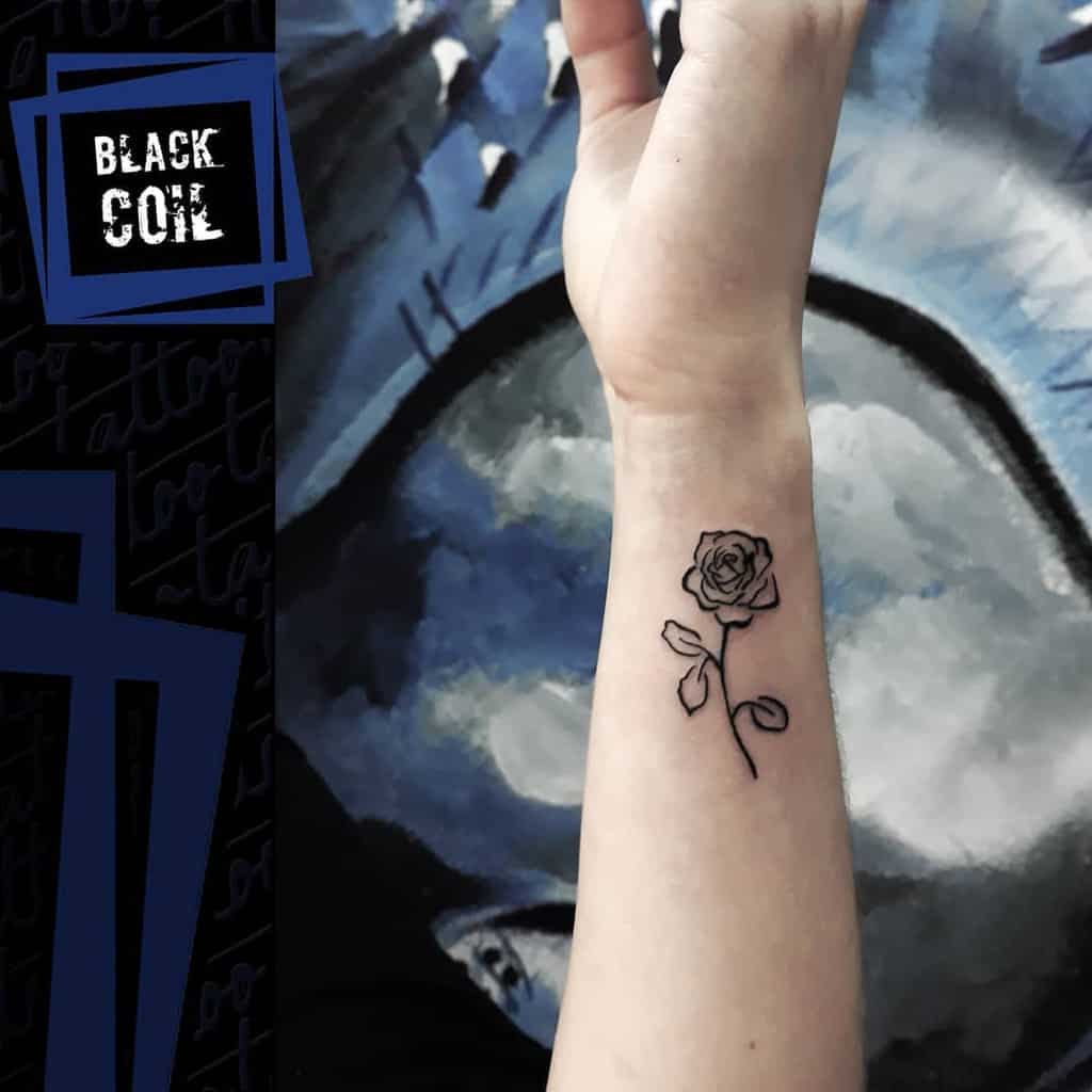 A small, minimalist black and white tattoo of a rose on the forearm, featuring delicate linework and a simple design