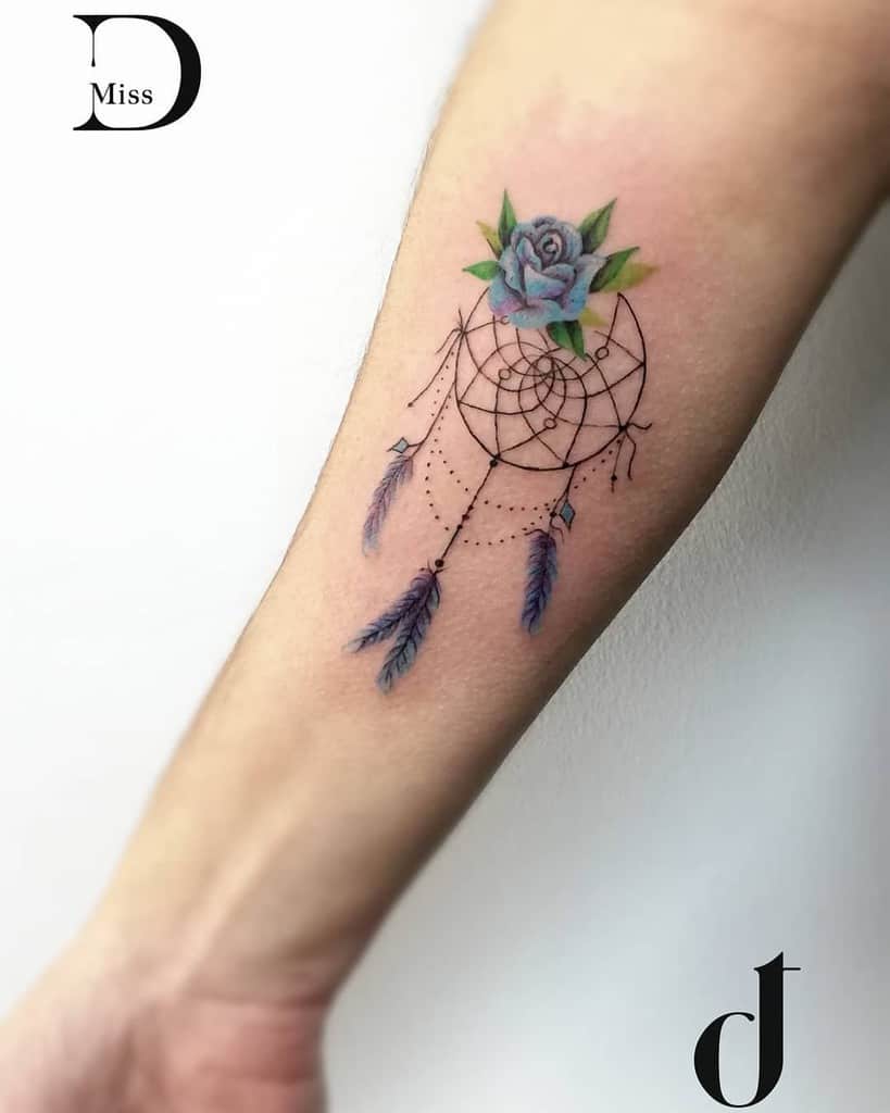 Forearm tattoo of a dreamcatcher with a blue rose and feathers