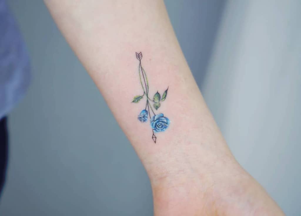 Tattoo of a bow and arrow with blue roses and green leaves on a person's arm