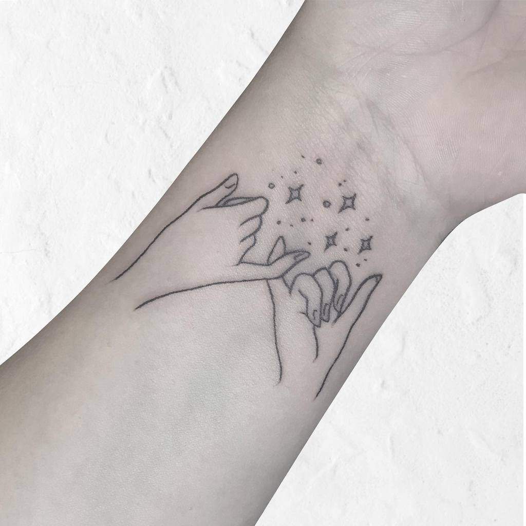 Tattoo of two hands making a pinky promise, surrounded by small stars and sparkles, on a wrist