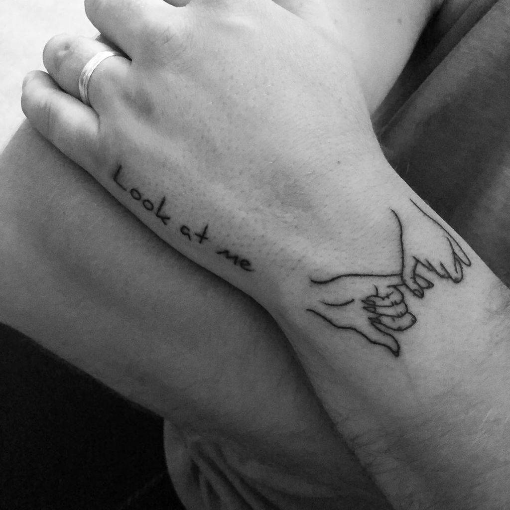 Minimalist pinky promise tattoo with fine black line art on the wrist, accompanied by the phrase "Look at me" in delicate handwriting