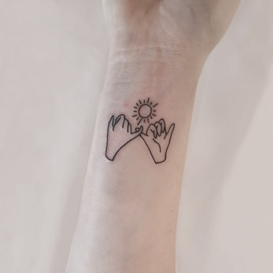 Tattoo of two hands making a pinky promise under a small sun, located on the inner wrist