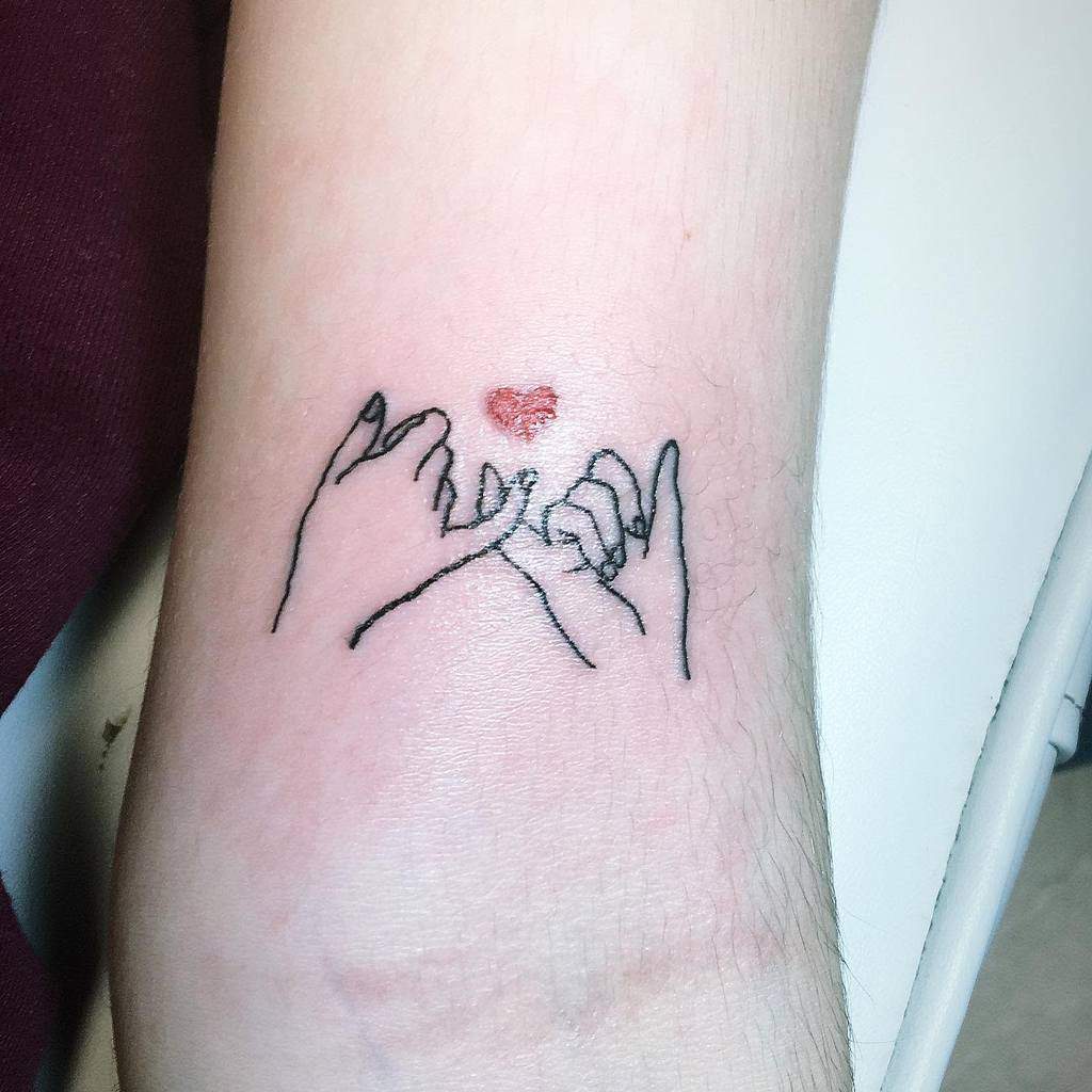Tattoo of two hands making a pinky promise with a small red heart above them on a forearm