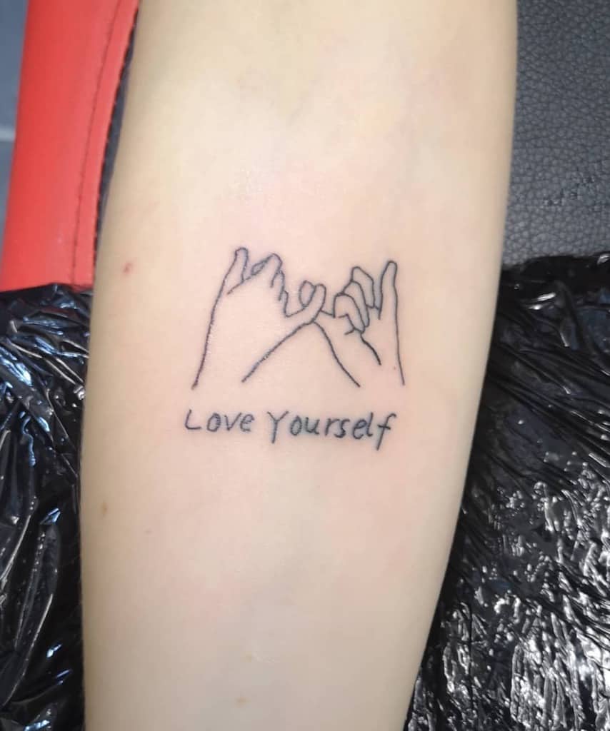 A tattoo of two hands linked by pinkies with the words "Love Yourself" below, on a forearm 