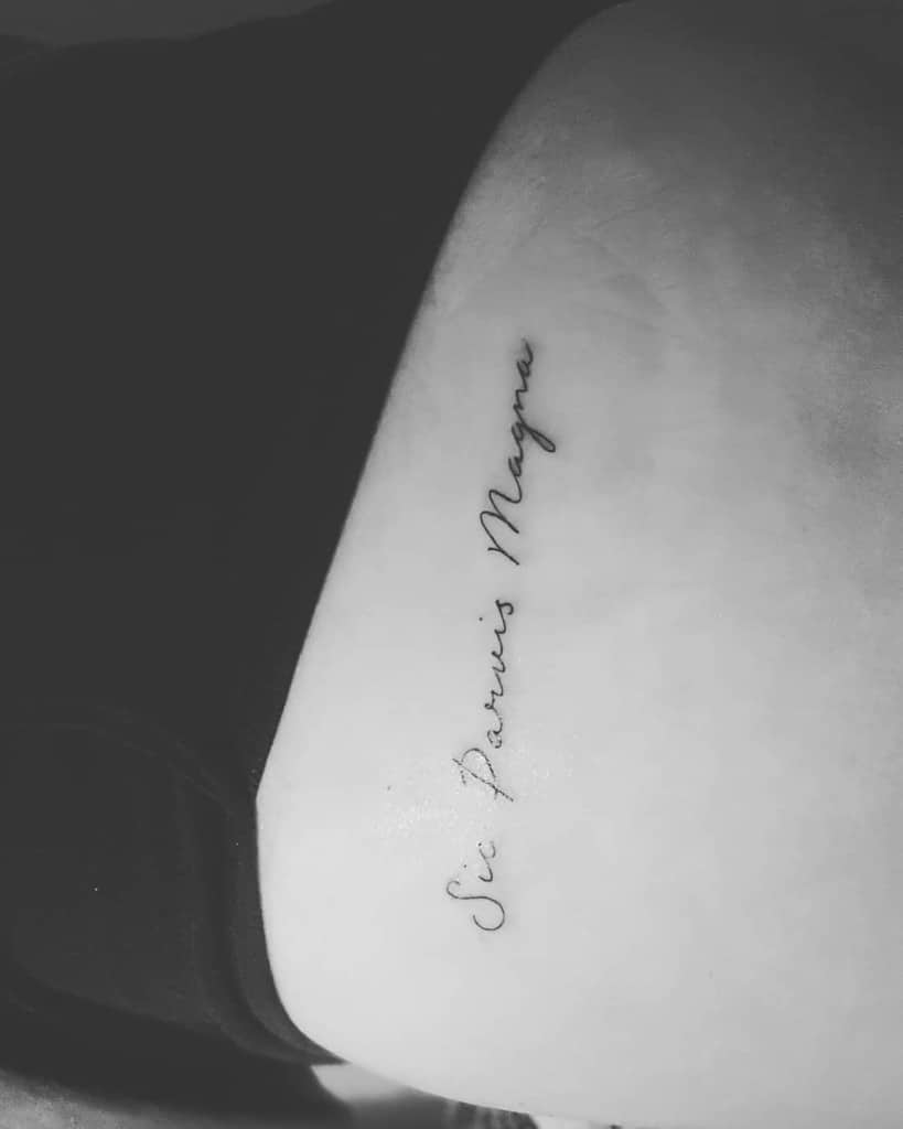 A tattoo of the phrase "Sic Parvis Magna" in cursive script on skin