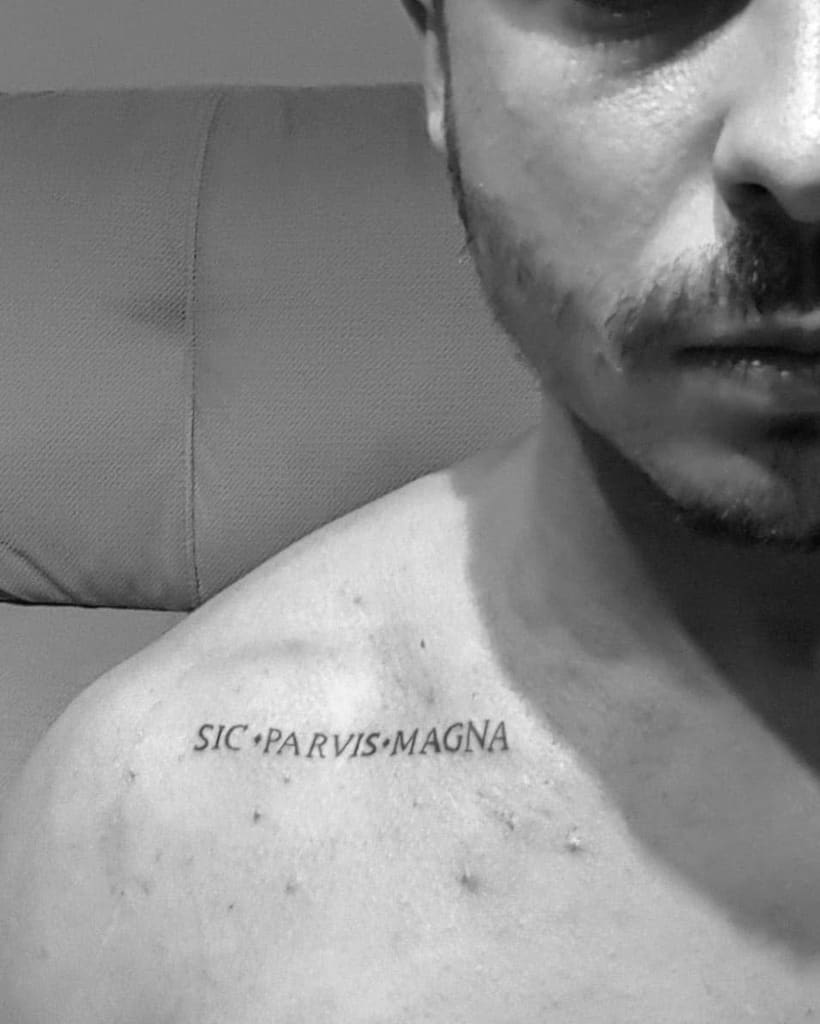 Man's shoulder tattoo with the phrase "SIC PARVIS MAGNA" in black ink