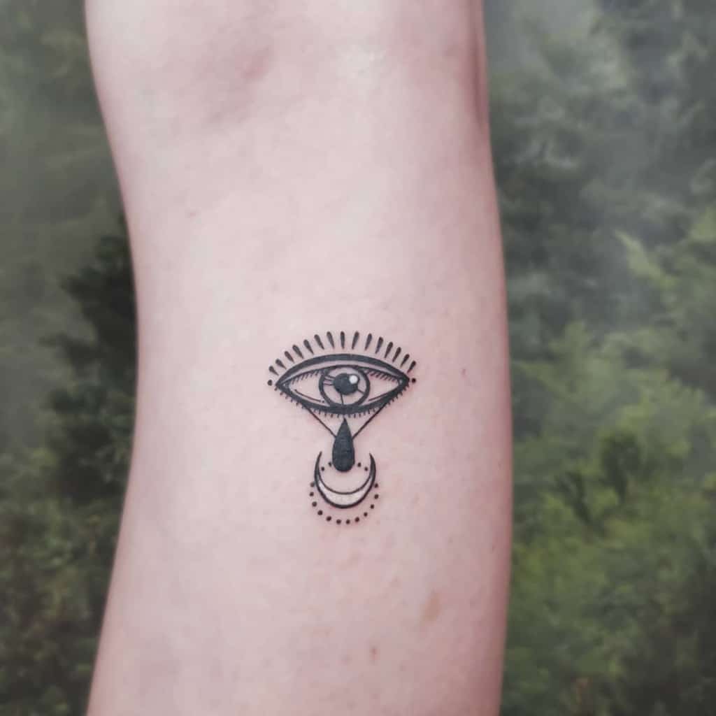 Minimalist tattoo of an all-seeing eye with a teardrop and a crescent moon, surrounded by radiating lines, inked in black on the arm.