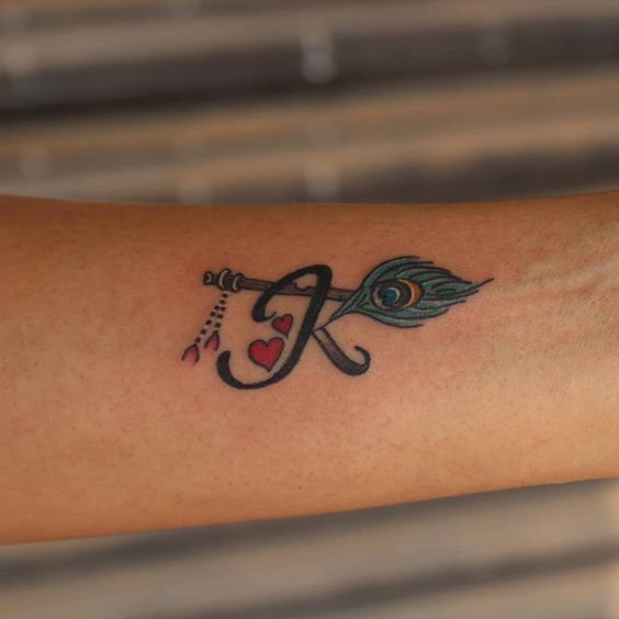 Small tattoo featuring a peacock feather attached to an arrow, intertwined with the letter 'K' and two red hearts, blending elegance with symbolism.
