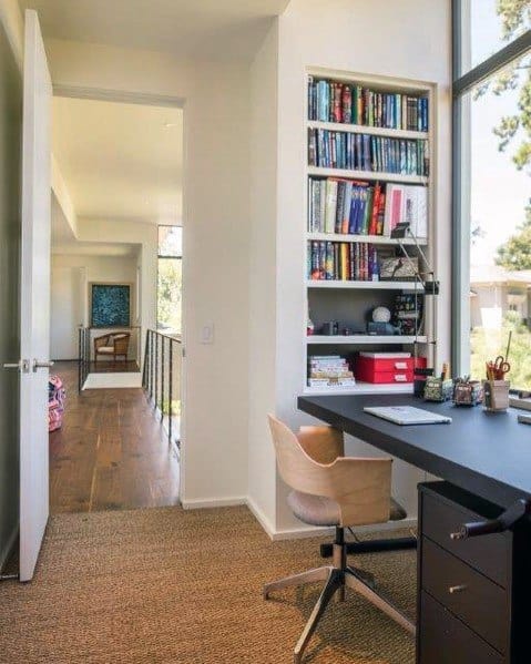 Compact home office with a built-in bookcase featuring colorful books and accessories, a sleek black desk, and a modern chair, complemented by a large window for natural light