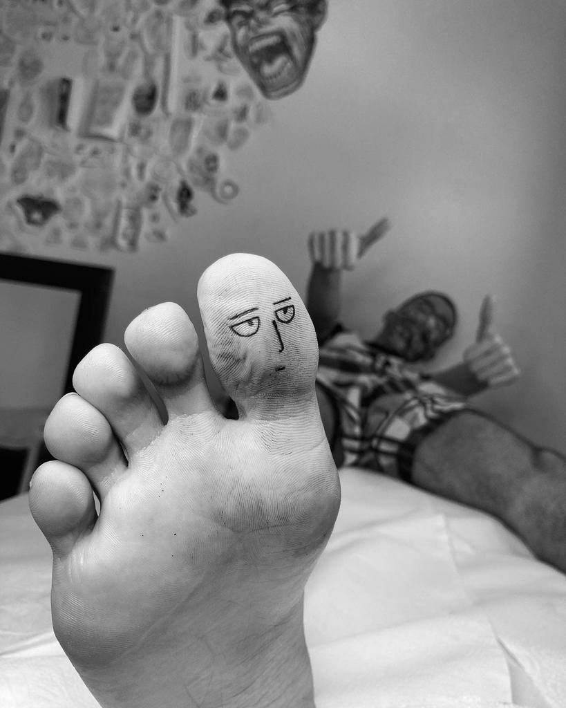 A creative and humorous tattoo of Saitama’s face from One Punch Man inked on a big toe, resembling his iconic deadpan expression