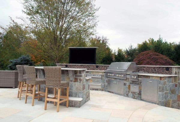 Stone outdoor BBQ grill bar with stainless steel appliances, wicker barstools, and a mounted TV.