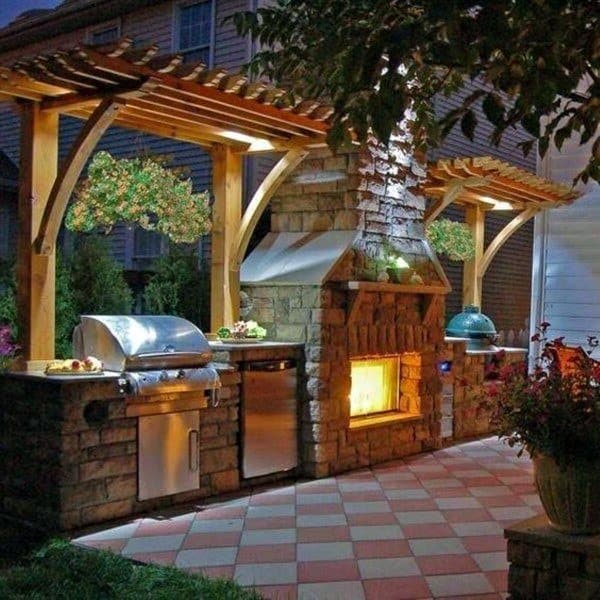 Outdoor kitchen with grill, oven, and fireplace under a pergola, beautifully illuminated at night with hanging plants