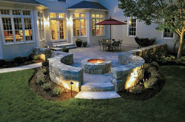 Charming patio with stone fire pit, seating, red umbrella, lush plants, and evening lighting