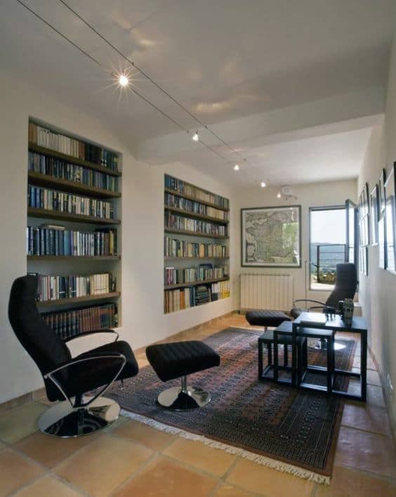 A cozy reading room with bookshelves, two black chairs, a rug, and a large window with a scenic view