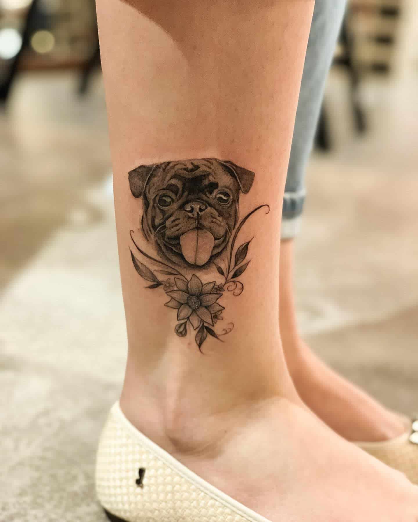 A detailed black-and-gray tattoo of a pug's face with its tongue out, surrounded by decorative floral elements and leaves