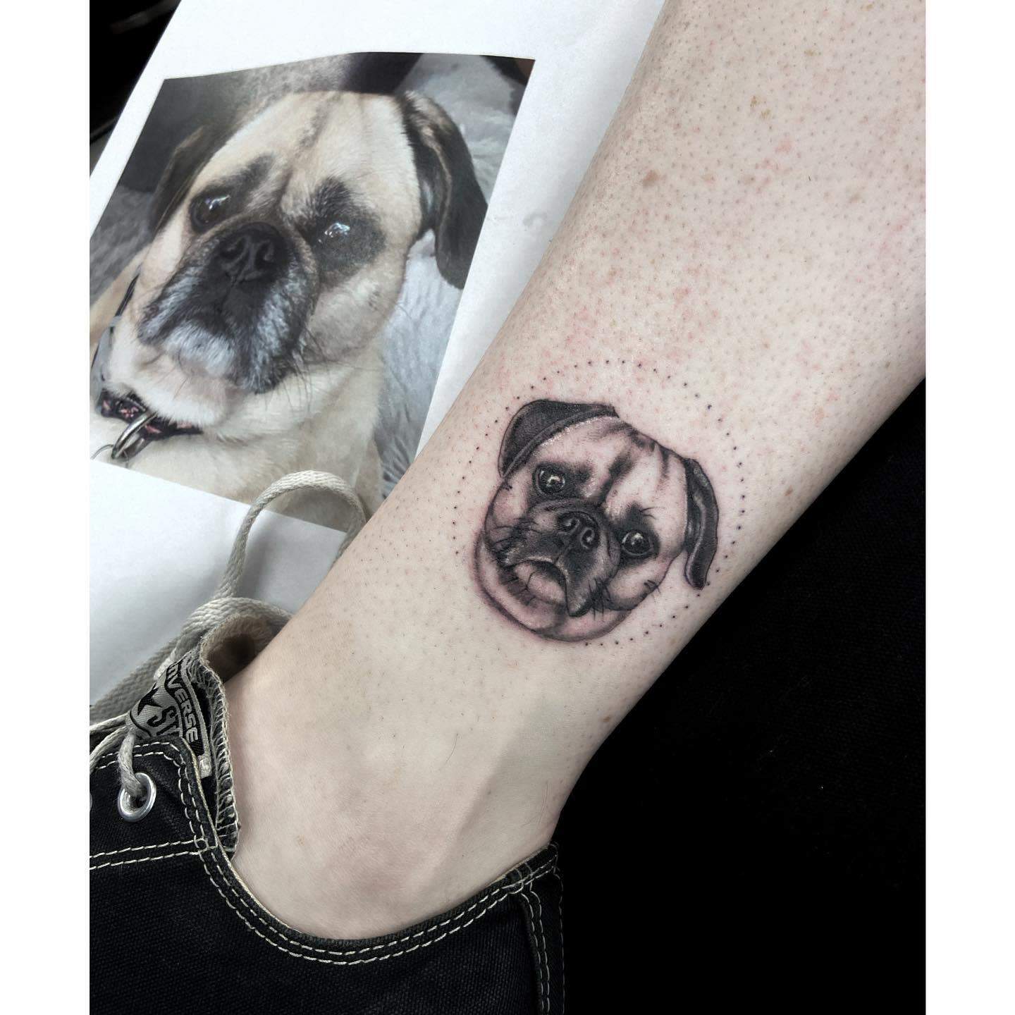 A realistic black-and-gray tattoo of a pug's face inside a dotted circular frame, with soft shading and fine details