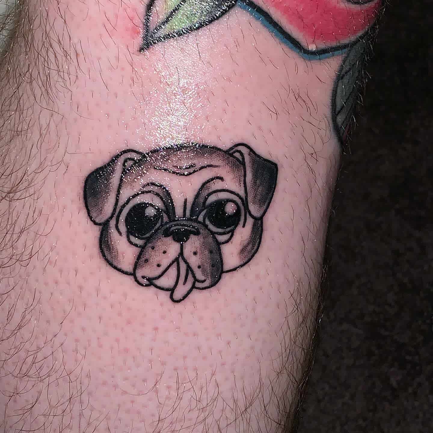 A cute black-and-gray tattoo of a pug's face with large expressive eyes and its tongue sticking out
