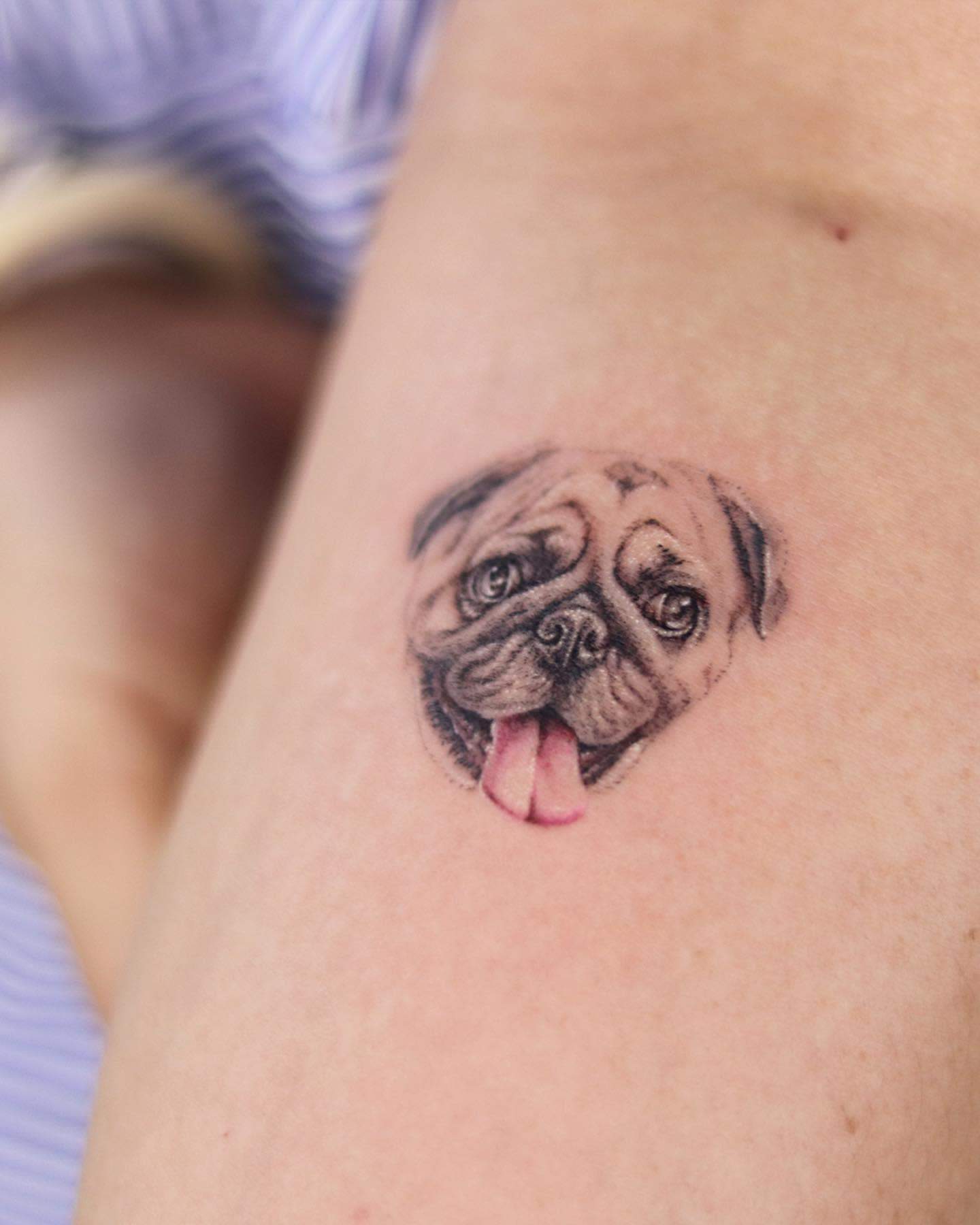 A highly detailed tattoo of a pug's face, capturing its expressive eyes and playful smile with its tongue sticking out