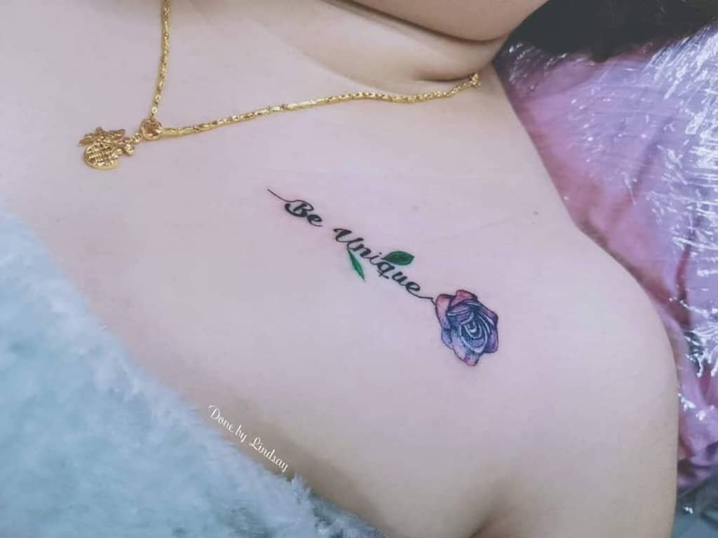 A small tattoo featuring the phrase "Be Unique" with a delicate purple rose beneath it, placed near the collarbone