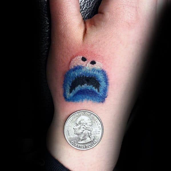 Tiny Cookie Monster tattoo on the wrist with a surprised expression, shown next to a quarter for scale.