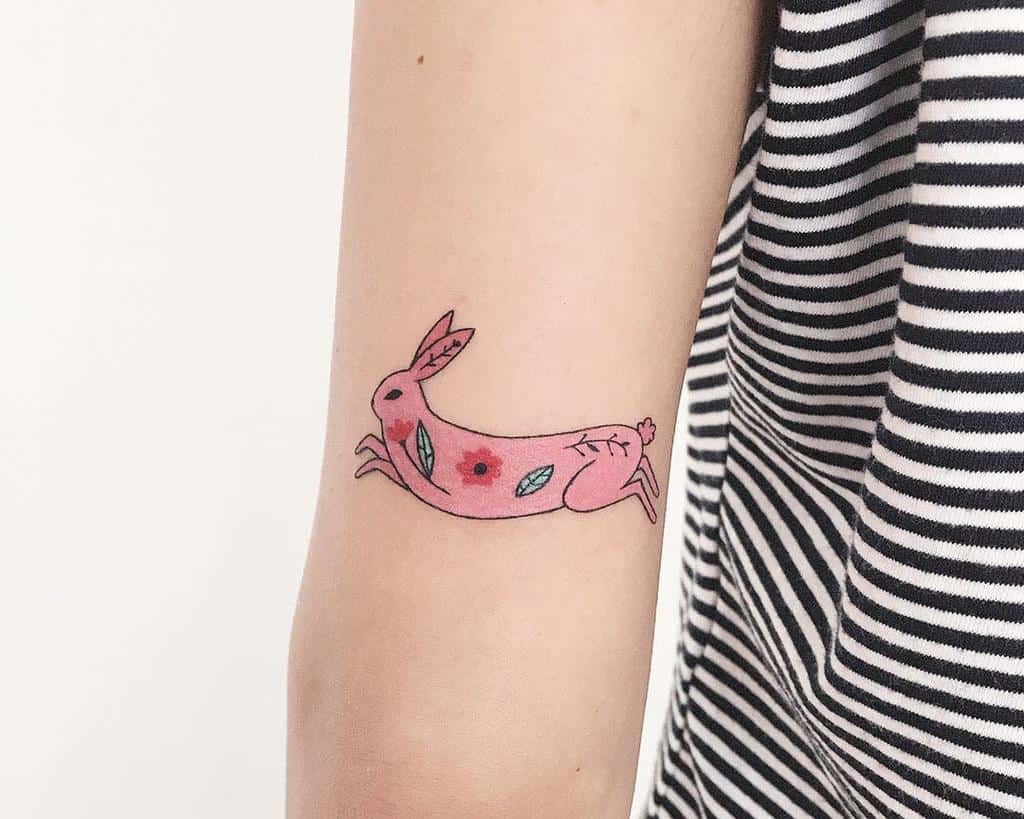 Whimsical tattoo of a pink rabbit in motion, adorned with colorful flowers and leaves on its body