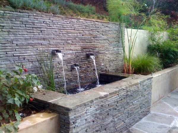 Stacked stone retaining wall with water spouts, a small pond, and decorative plants.