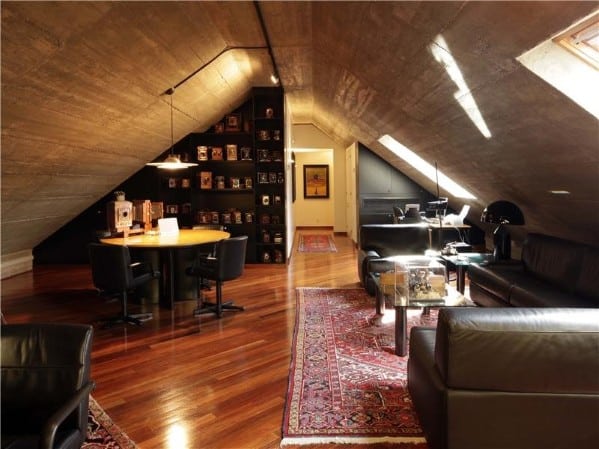 Modern attic with leather couches, round table, bookshelves, and sloped ceiling; warm lighting and wooden floors